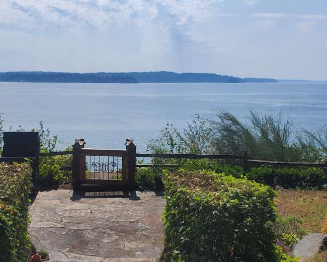 The image shows a stone patio with a wrought iron gate leading to a view of a calm waterfront scene surrounded by greenery.