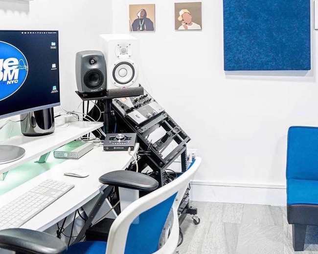 The image shows a modern music studio with a computer setup, speakers, and soundproofing panels on the walls.
