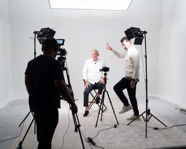A man sits on a stool in a brightly lit studio while another man jumps playfully nearby, surrounded by cameras and lights.