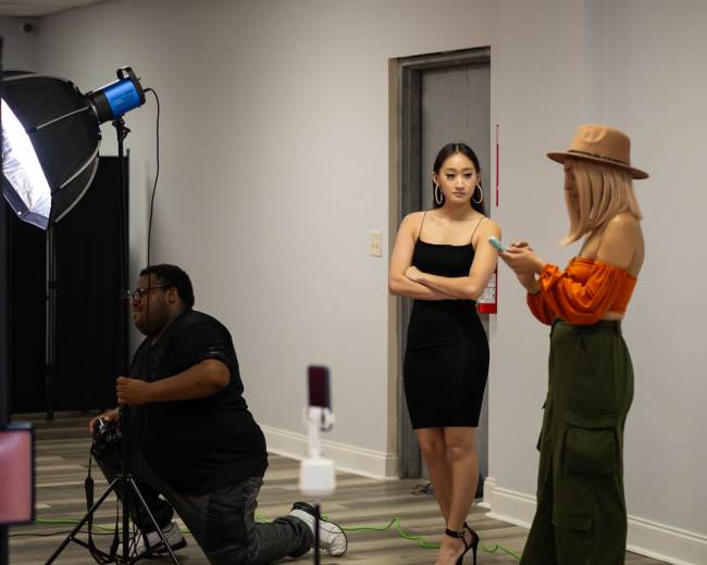 A photographer sets up a lighting equipment while two women pose and check their phones in a studio environment.