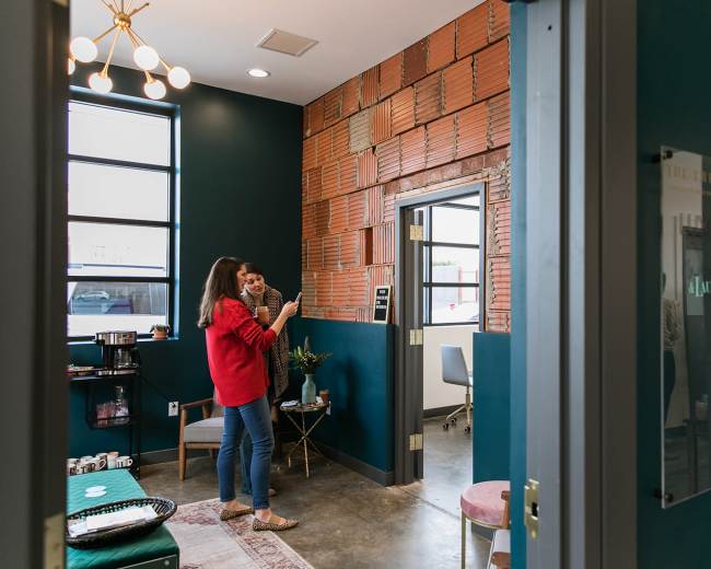 A woman in a red sweater talks to another person while standing in a room with green walls and exposed brick accents.