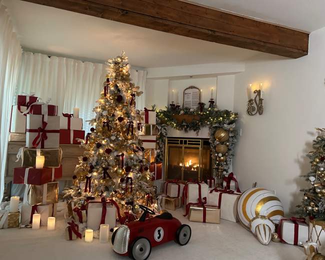 The image shows a decorated living room with a Christmas tree surrounded by wrapped gifts, a fireplace with candles, and a vintage toy car in the foreground.