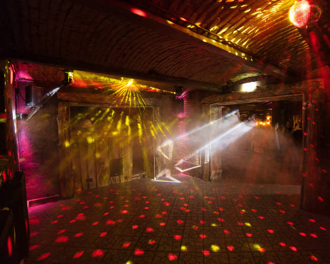 The interior of a dimly lit venue features colorful light beams and a disco ball casting patterns across the floor and walls.