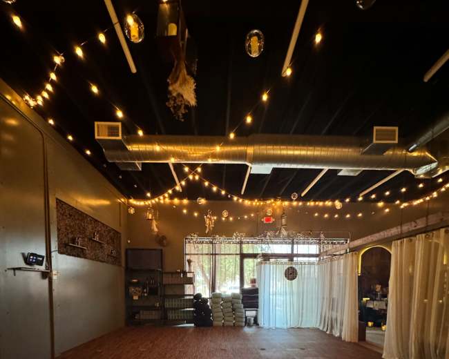 The image shows a spacious indoor studio with wooden flooring, draped curtains, and soft lighting from hanging bulbs.