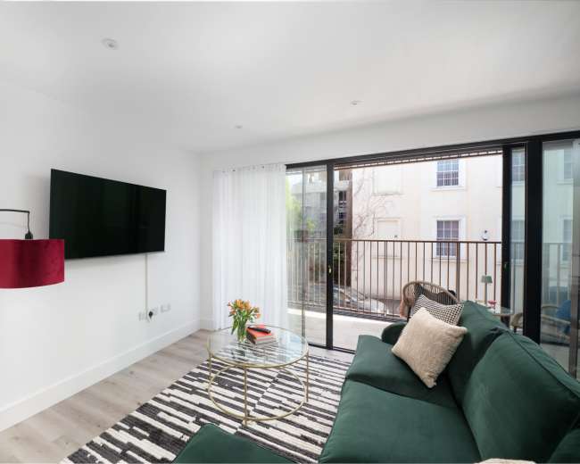 A modern living room features a green sofa, a round glass table, a large wall-mounted television, and sliding glass doors opening to a balcony.