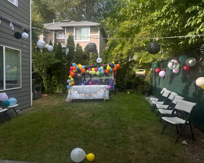 The image shows a decorated backyard with a colorful balloon arch, a festive table covered with a tablecloth, and white chairs arranged for a celebration.