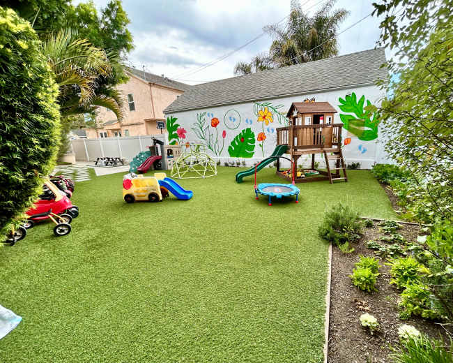 The image depicts a vibrant backyard play area featuring a wooden play structure, a small slide, a trampoline, and various colorful ride-on toys, all situated on artificial grass with a mural-covered wall in the background.