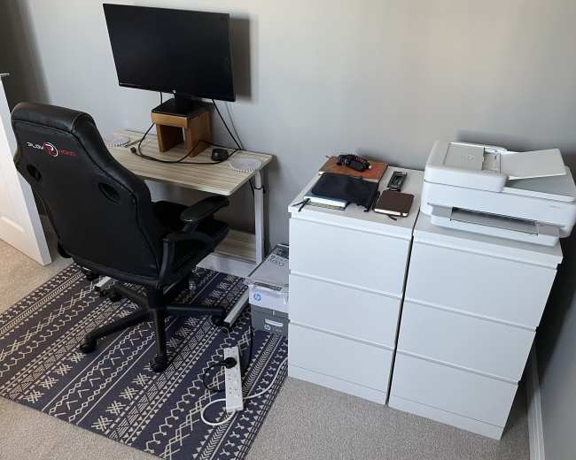 A small home office setup features a desk with a monitor and chair, alongside a printer and storage cabinets.