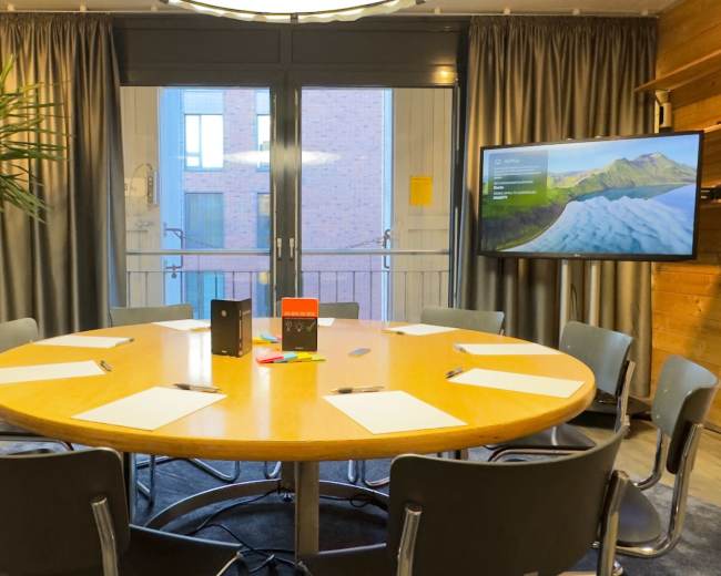 A round wooden table surrounded by eight chairs is set up in a conference room with large windows and a screen displaying a landscape image.