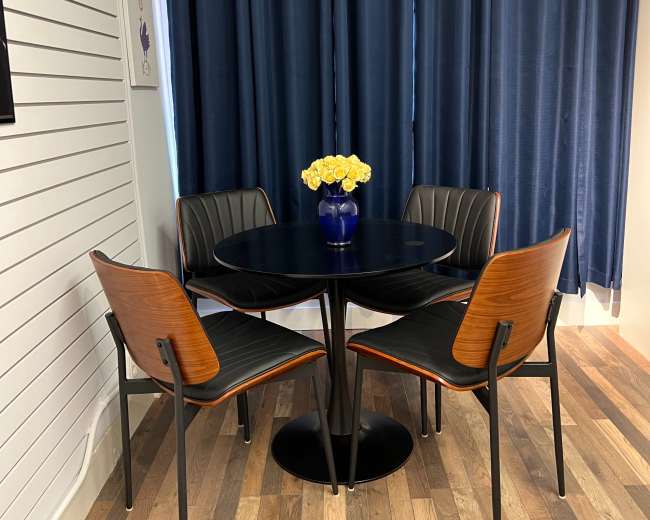 A round black dining table with four wooden chairs is positioned in front of a blue curtain, complemented by a vase of yellow flowers on the table.