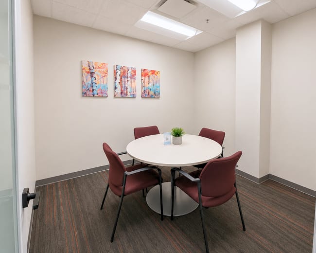 The image shows a small conference room with a round table surrounded by four maroon chairs and three colorful abstract paintings on the wall.