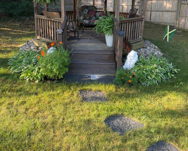 A wooden gazebo with a hanging chair and potted plants stands in a green yard surrounded by flowers and stones, with a fence and trees in the background.