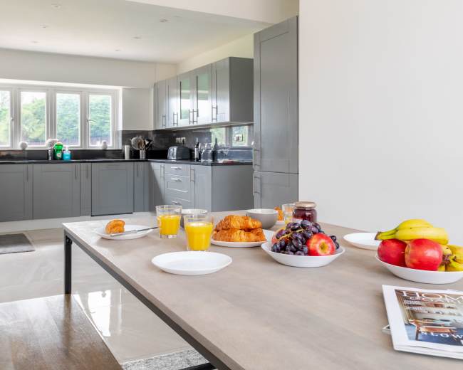 A modern kitchen features a wooden table set with a variety of breakfast items, including pastries, fruit, and beverages, while sleek gray cabinetry lines the walls.