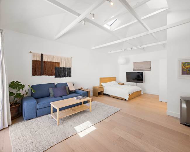 The image shows a minimalist bedroom with a blue sofa, a wooden bed, a television mounted on the wall, and a large window allowing natural light to enter.