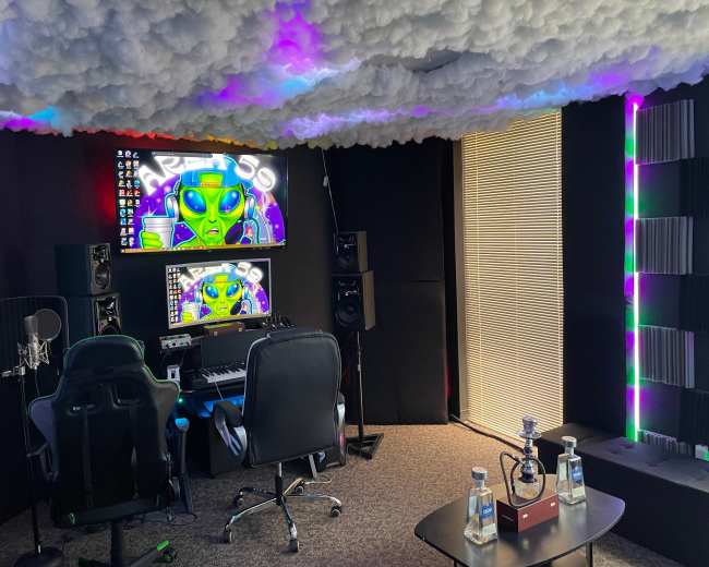 The image shows a music studio featuring a desk with a digital audio workstation, two monitors displaying colorful graphics, a microphone setup, and colorful lighting against a textured ceiling.