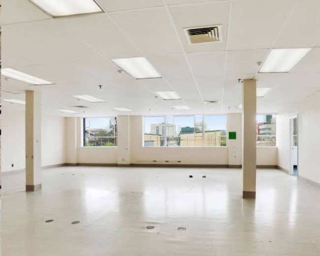 The image shows a spacious, empty room with white walls, fluorescent lighting, and a window that provides a view of the outdoors.