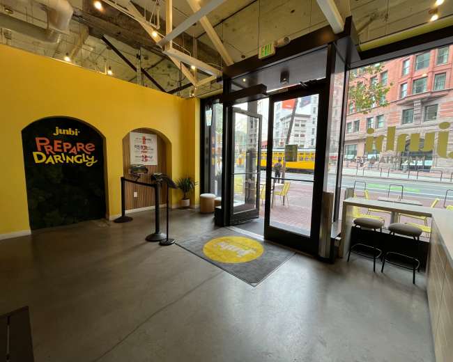The image shows the interior of a café with a bright yellow wall, a welcoming entrance, and seating areas visible through large glass doors.