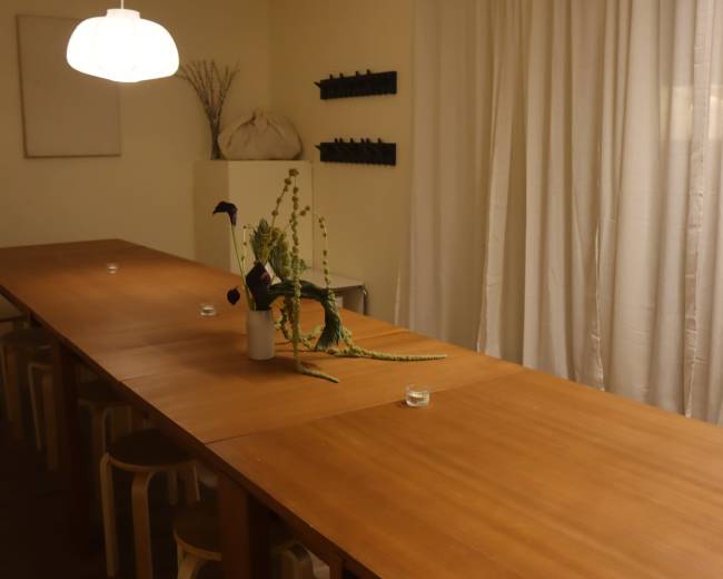 A long wooden table with several chairs is set in a softly lit room featuring white curtains and simple decor.