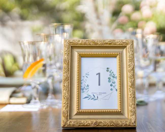 A gold-framed table number display is set on a wooden table, surrounded by glassware and decorative elements, indicating the number one.