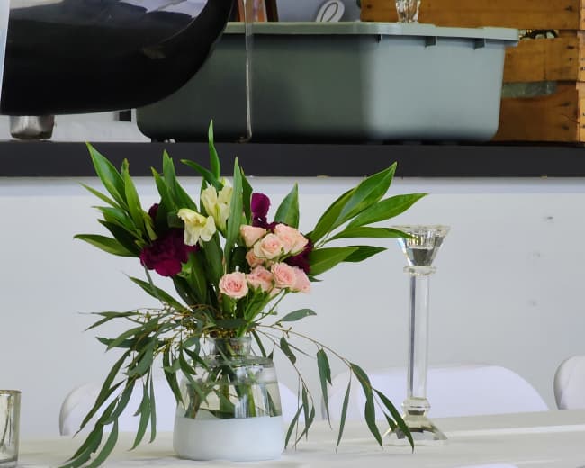 A simple floral arrangement is placed in a glass vase on a table set with white chairs and a white tablecloth.
