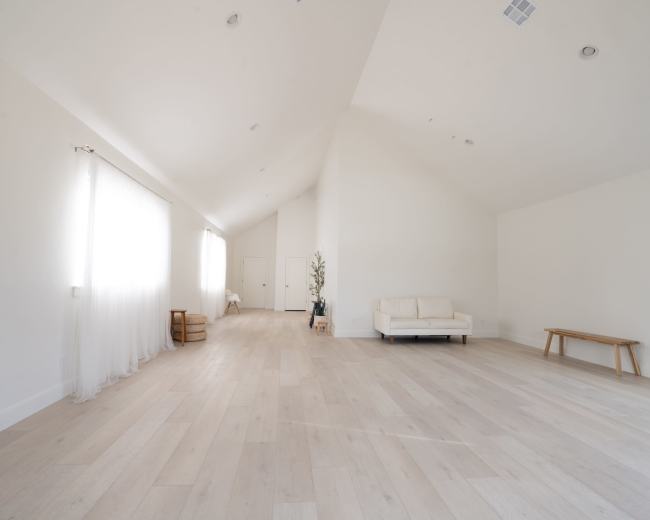 The image shows a bright, spacious room with light-colored wooden flooring, large windows, and minimal furnishings including a white sofa and a wooden bench.
