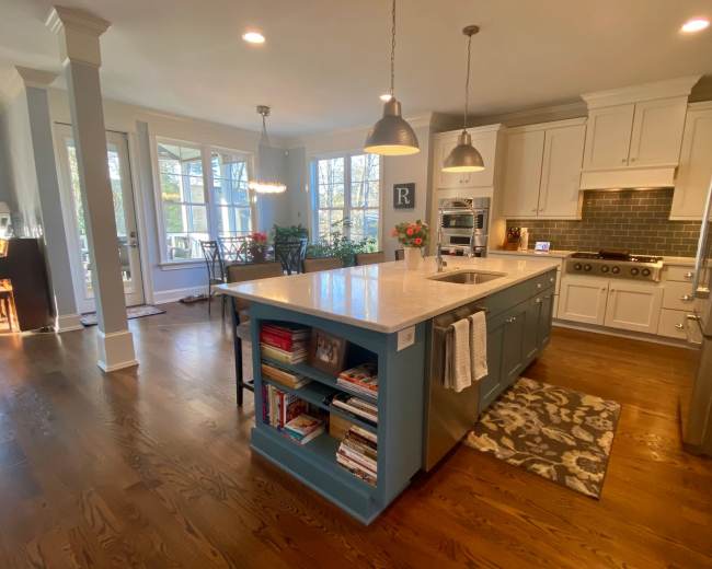 The image shows a modern kitchen featuring a large central island with blue cabinetry, white countertops, and stainless steel appliances, illuminated by overhead pendant lights.