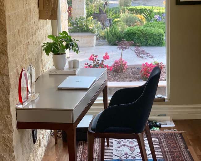 A modern desk and chair are positioned by a large window, looking out onto a landscaped garden with flowers and greenery.