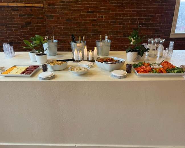 A food spread is arranged on a white table with platters of cheese, charcuterie, vegetables, and chips, alongside drinks in ice buckets and candles.