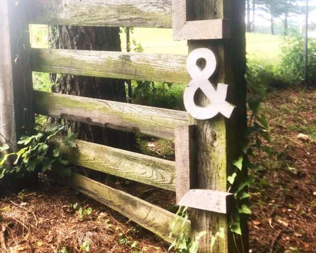 A weathered wooden gate with an ampersand symbol affixed to its side, surrounded by greenery.