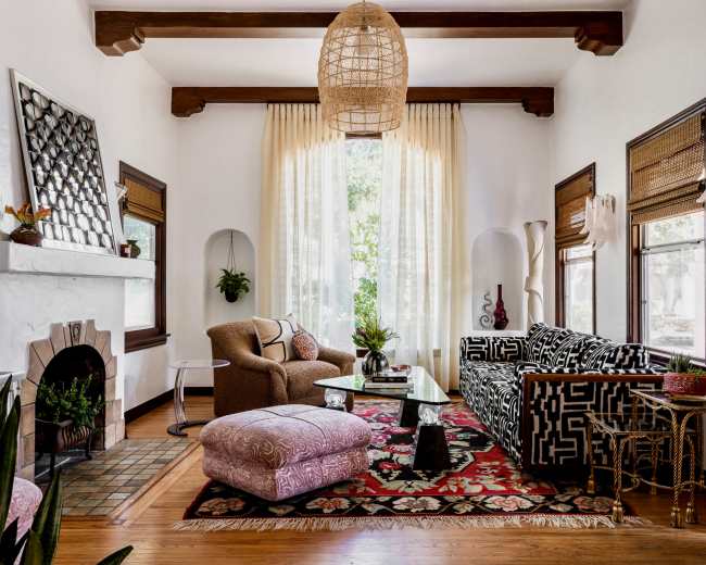 The scene depicts a cozy living room with a patterned black and white sofa, a relaxed chair, a coffee table, and an area rug, all complemented by large windows and light-colored drapes.