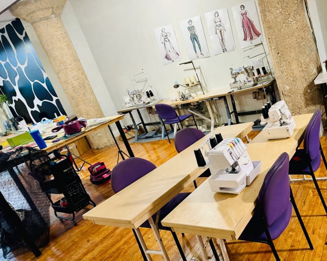 The image shows a sewing studio with several workstations equipped with sewing machines and tables, along with fashion sketches displayed on the wall.