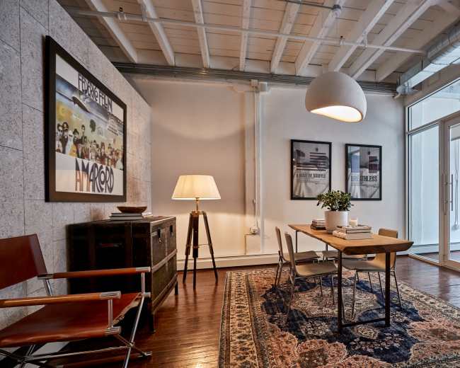 The image shows a modern workspace with a table and chairs, framed artwork on the wall, a floor lamp, and a patterned rug on hardwood flooring.