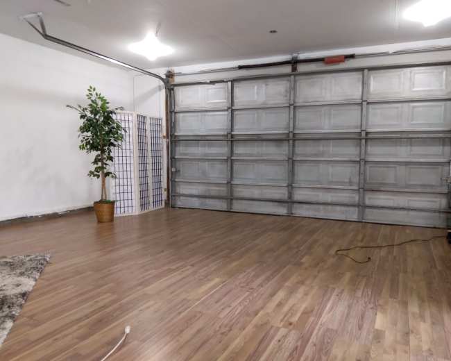 An empty garage features a large metal door, a potted plant in the corner, and wooden flooring.