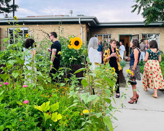 A group of people gathers outside a building, surrounded by vibrant gardens filled with flowers and greenery.