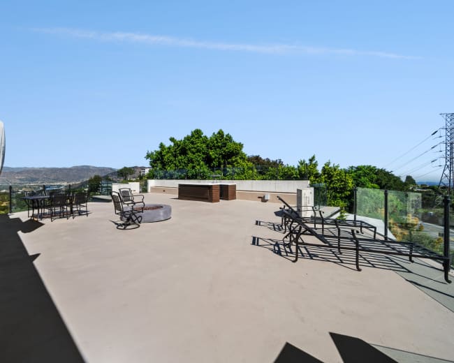 A spacious rooftop terrace with metal furniture and a fire pit, surrounded by glass railings and greenery in the background.