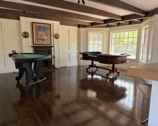 The image shows a room with wooden beams, polished floors, and a poker table alongside a pool table, illuminated by natural light from large windows.