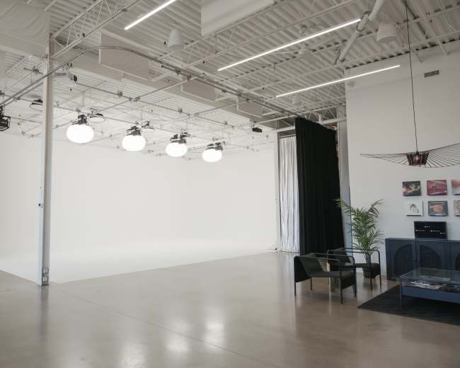 The image shows a spacious, well-lit studio with a plain white backdrop, equipped with overhead lighting and minimal furniture.