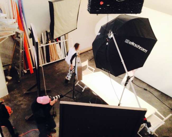 A photography studio is set up with lighting equipment, a backdrop, and a model posing at a table.