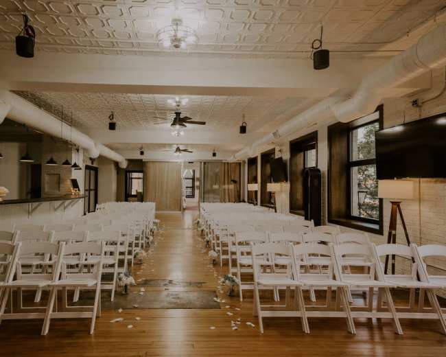 An indoor wedding venue features rows of white chairs arranged facing a central aisle, with overhead fans and pendant lights.