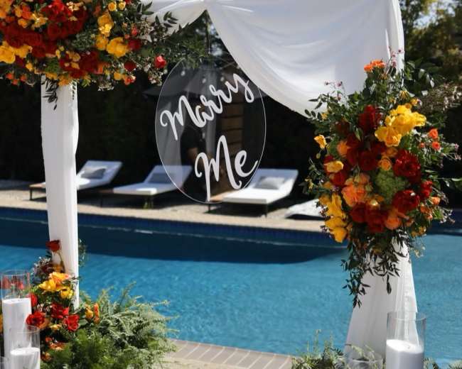A decorated arch with vibrant flowers and a "Marry Me" sign overlooks a swimming pool surrounded by lounge chairs.