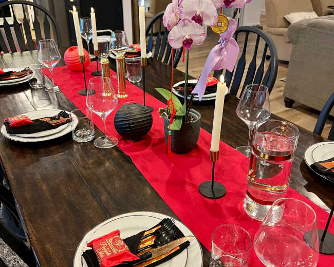 A dining table is set with plates, glasses, and silverware, featuring a red table runner adorned with an orchid centerpiece and candles.