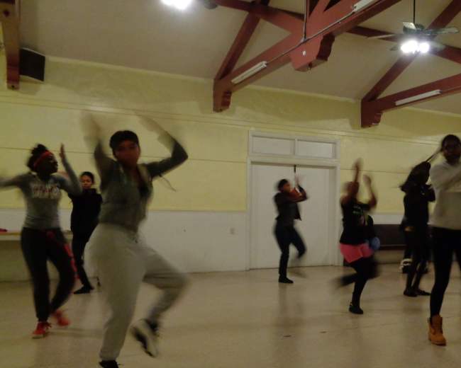 A group of people is dancing in a spacious room with wooden beams and overhead lights.