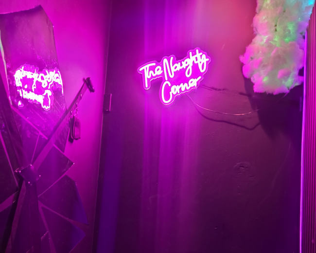 The image shows a dimly lit corner with a neon sign that reads "The Naughty Corner," surrounded by purple lighting and a partially opened umbrella.