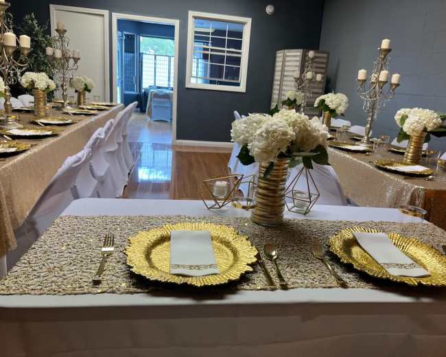 A decorated dining area features tables set with gold tableware, white floral centerpieces, and elegant candle holders.