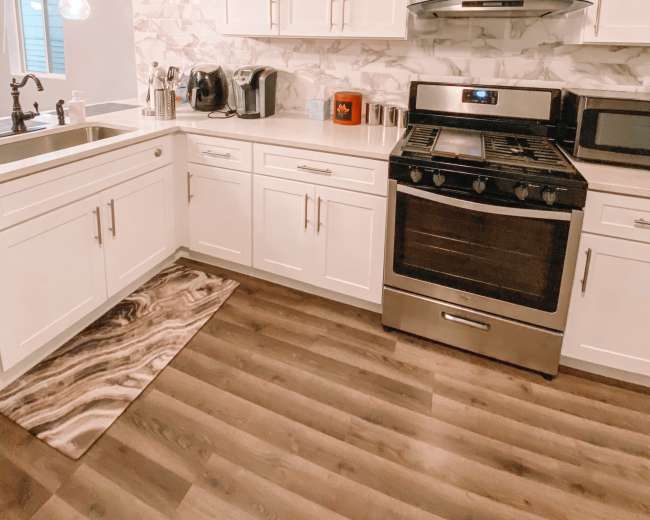 The image shows a modern kitchen with white cabinets, a stainless steel stove, and a patterned rug on the wooden floor.