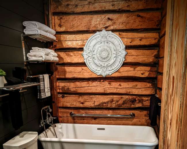 The image shows a rustic bathroom featuring wooden walls, a white bathtub, a toilet, a sink, and decorative elements including towels and a wall ornament.