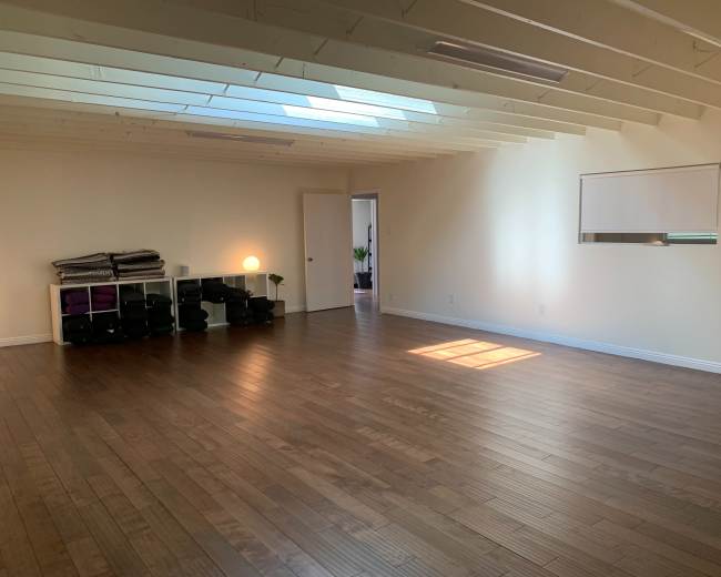 The image shows a spacious, empty room with wooden floors, a large window letting in light, and a shelf containing yoga mats and blocks.
