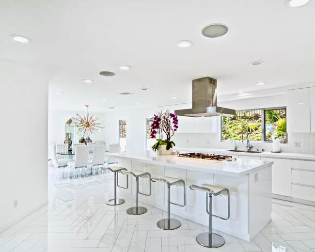 The image shows a modern kitchen with sleek white cabinetry, a large island with bar stools, and an adjoining dining area visible through large windows.