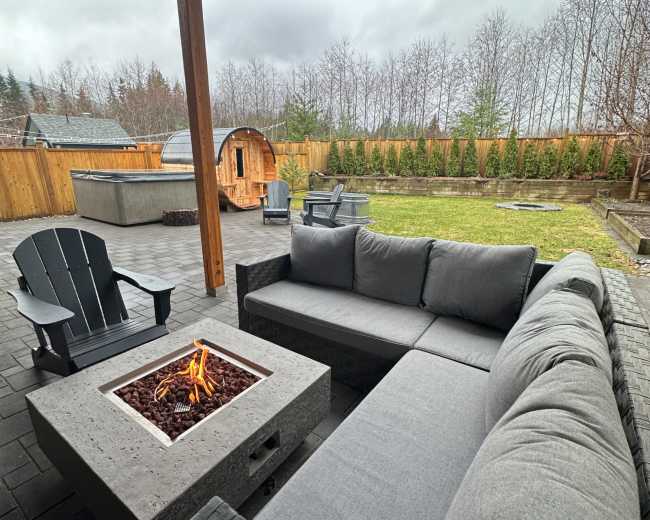 A modern outdoor seating area features a gray sectional sofa and a square fire pit, surrounded by a grassy yard and wooden fencing under a cloudy sky.