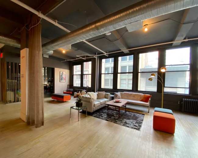The image shows a spacious modern loft with large windows, wooden floors, and a minimalist furniture arrangement, including a couch, coffee table, and colorful ottomans.